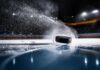A black hockey puck sliding across an ice rink with ice particles flying around it