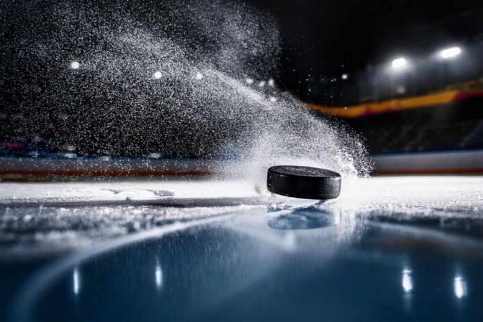A black hockey puck sliding across an ice rink with ice particles flying around it