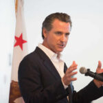 A man in a suit speaking into a microphone with a California flag in the background