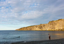 A man standing on a rocky beach near the water with cliffs in the background
