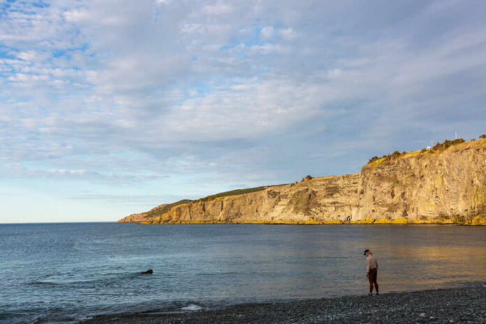 A man standing on a rocky beach near the water with cliffs in the background