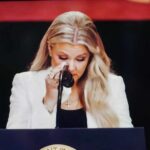 A woman in formal attire wiping her tears while speaking at a podium