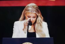 A woman in formal attire wiping her tears while speaking at a podium