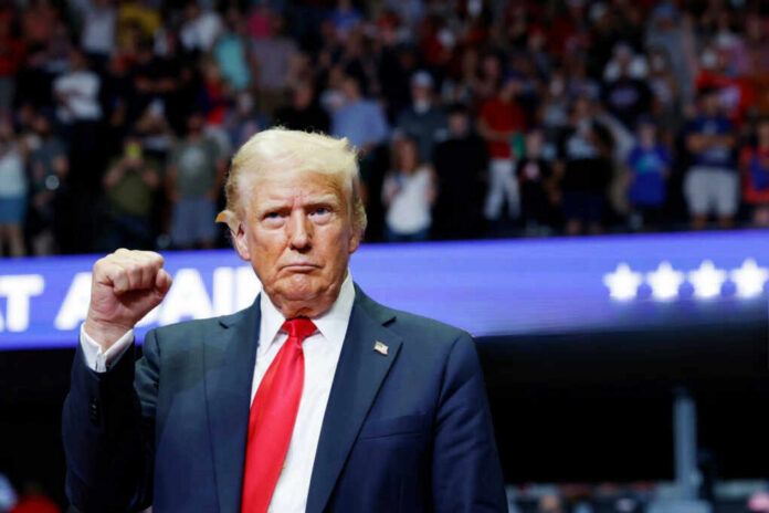 A man in a suit with a red tie raises his fist at a rally, with a cheering crowd in the background