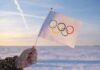 A hand holding an Olympic flag against a snowy landscape during sunset