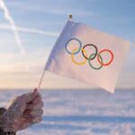 A hand holding an Olympic flag against a snowy landscape during sunset