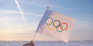 A hand holding an Olympic flag against a snowy landscape during sunset