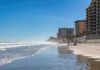 A beach with people walking along the shore and buildings in the background under a clear blue sky