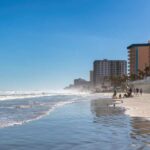 A beach with people walking along the shore and buildings in the background under a clear blue sky