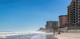 A beach with people walking along the shore and buildings in the background under a clear blue sky