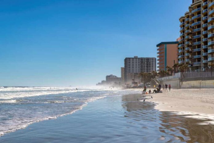 A beach with people walking along the shore and buildings in the background under a clear blue sky