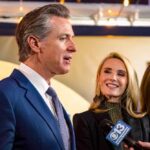 A man speaking at a public event with two women nearby, one smiling and another looking down