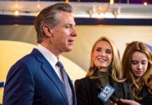 Governor’s Wife Sparks Massive Conflict Scandal A man speaking at a public event with two women nearby, one smiling and another looking down