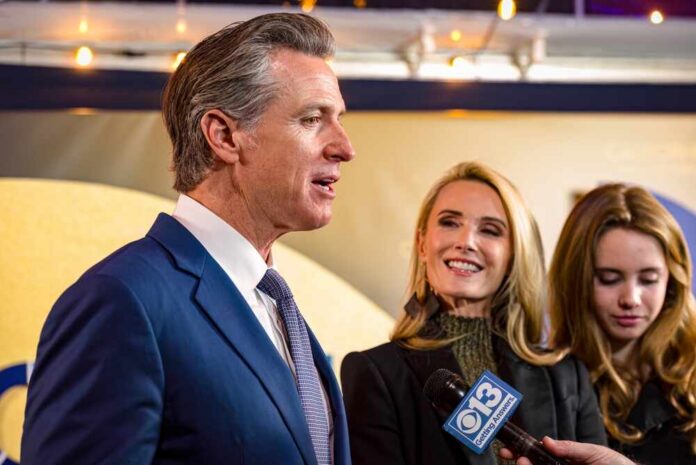 A man speaking at a public event with two women nearby, one smiling and another looking down