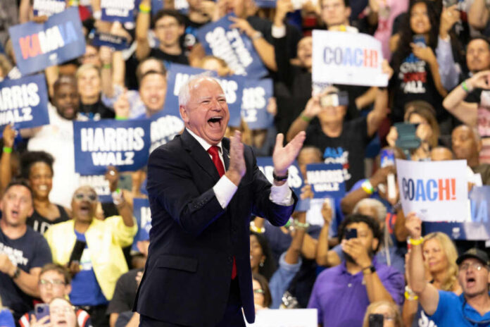 A man in a suit enthusiastically claps at a political rally with a cheering crowd holding signs