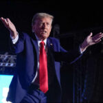 A man in a blue suit and red tie gestures while speaking at a nighttime rally