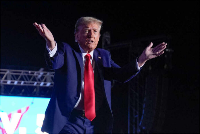 A man in a blue suit and red tie gestures while speaking at a nighttime rally