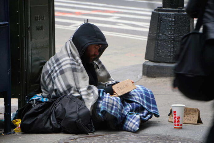 A homeless man sitting on the street wrapped in a blanket with a sign asking for help