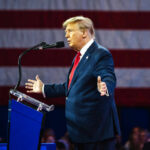 A man in a suit speaking at a podium with an American flag in the background