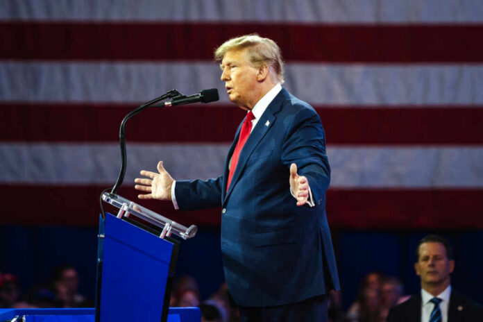 A man in a suit speaking at a podium with an American flag in the background