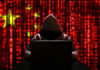 A hooded figure sitting in front of a laptop with a red digital background