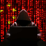 Hacker Bust: COVID-19 Secrets Stolen by China A hooded figure sitting in front of a laptop with a red digital background