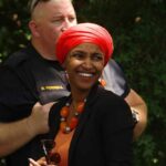 Smiling woman in a red headscarf at a community event with a police officer in the background