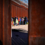 A line of people waiting near a border wall with barbed wire