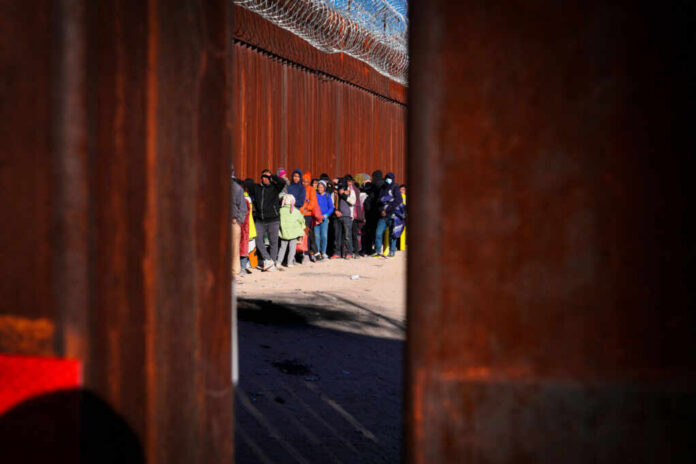 shutterstock_2241078561.jpg A line of people waiting near a border wall with barbed wire