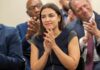 AOC Teams Up With Controversial 9/11 Commentator A woman in a dark dress clapping at a political event with an engaged audience in the background