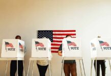 Individuals casting their votes in a polling station with an American flag in the background