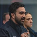 A man in a mayor's jacket speaking at a press conference with a woman in the background