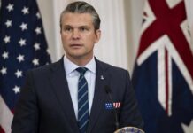 A man in a suit speaking at a podium with flags in the background