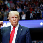 Trump’s CEO Entourage Sparks Controversy A man in a suit with a red tie raises his fist at a rally, with a cheering crowd in the background