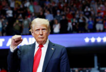 A man in a suit with a red tie raises his fist at a rally, with a cheering crowd in the background