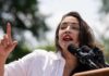 A woman passionately speaking at a rally with microphones in front of her