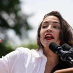 A woman passionately speaking at a rally with microphones in front of her