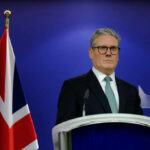 A political leader standing at a podium with a British flag in the background