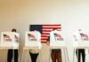 Individuals casting their votes in a polling station with an American flag in the background