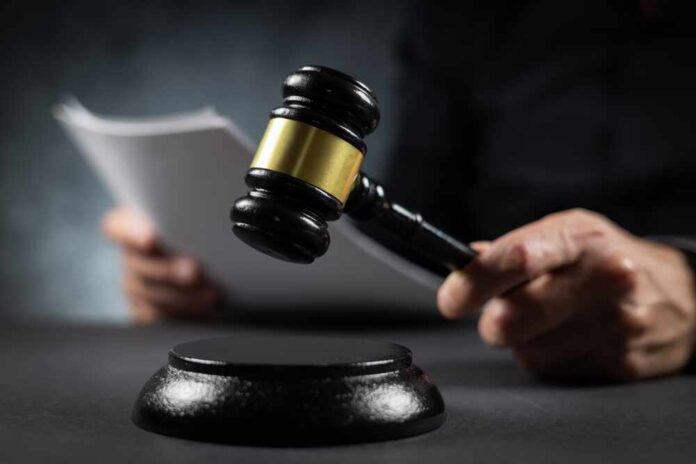 Judge,With,Paper,Document,Pronouncing,Sentence,In,A,Court,Of A gavel held above a sounding block with a person reading documents in the background
