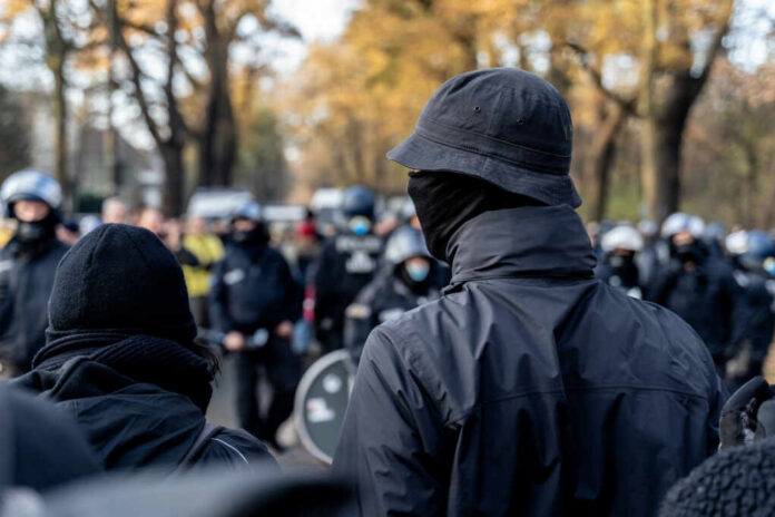 A crowd of masked individuals facing law enforcement in an autumn setting