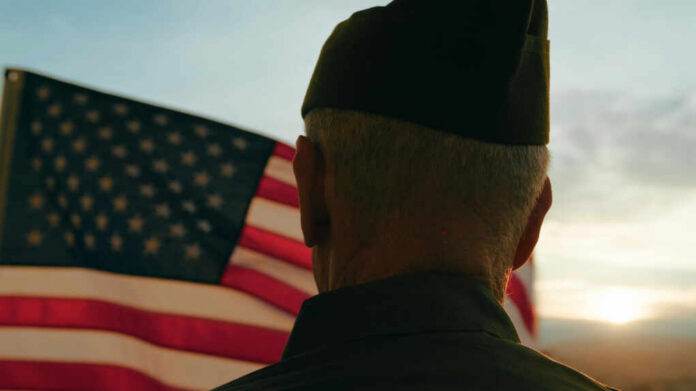 shutterstock_2513309275 A veteran in uniform standing in front of an American flag during sunset