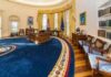 Oval office with wooden desk, blue rug, and gold curtains