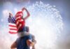 Trump’s Mega Boom Outsizes The World Child on adult's shoulders holding American flag during fireworks