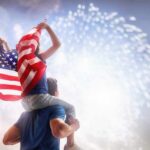 Child on adult's shoulders holding American flag during fireworks
