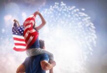 Trump’s Mega Boom Outsizes The World Child on adult's shoulders holding American flag during fireworks