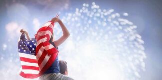 Child on adult's shoulders holding American flag during fireworks