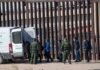 Border agents escort a group near a tall fence and a patrol van