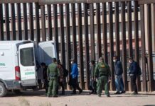 Border agents escort a group near a tall fence and a patrol van