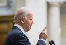 Joe Biden speaking at a podium, pointing upward with his index finger